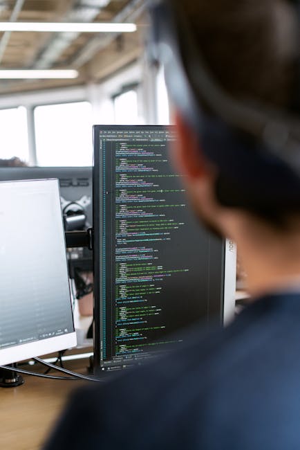 The image shows a person working at a desk with multiple computer monitors. One screen displays lines of colorful code on a dark background, indicating programming or web development work. The workspa